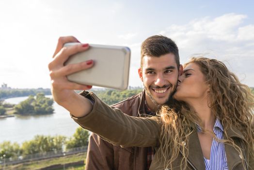 Beautiful young woman taking selfie in the nature while kissing her handsome boyfriend