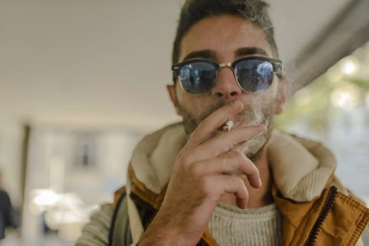 Young handsome hipster with orange jacket and sunglasses enjoying a cigarette in the street.