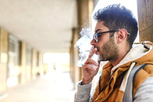 Young handsome hipster with orange jacket and sunglasses enjoying a cigarette in the street.