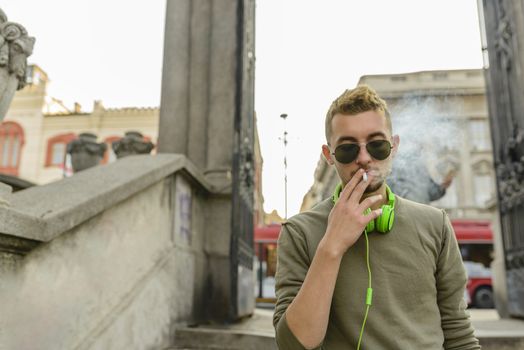 Young handsome man with green headphones and sunglasses enjoying a cigarette in the street.