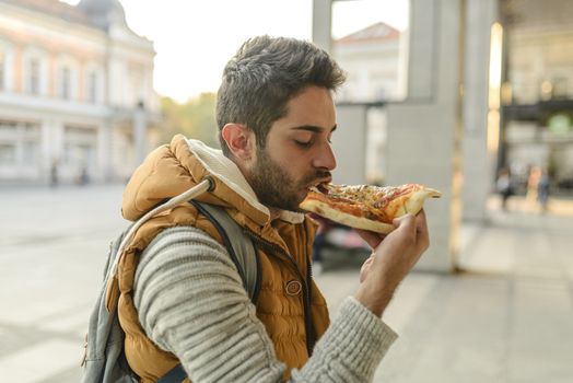 Handsome bearded young man with orange jacket eating pizza in the city street