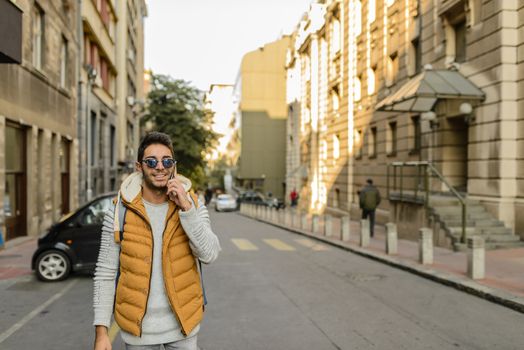 Young hipster with orange jacket, sunglasses, backpack and his smart phone in the city street