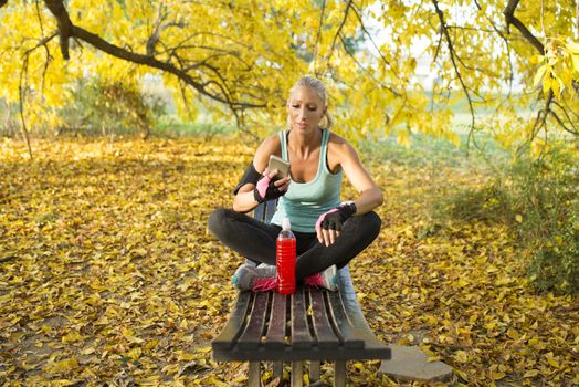 Attractive blonde sportswoman refreshing with cold drink using her smart phone. Sitting on the bench in the public park. Healthy lifestyle concept.