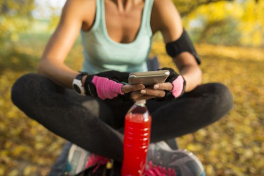 Attractive blonde sportswoman refreshing with cold drink using her smart phone. Sitting on the bench in the public park. Healthy lifestyle concept.