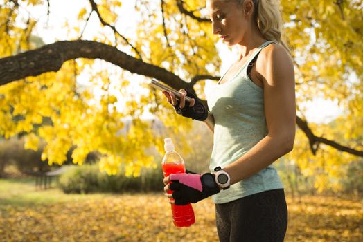 Attractive blonde sportswoman refreshing with cold drink and using her smart phone. Healthy lifestyle concept.
