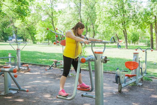 Pregnant woman is doing exercises outdoors on fitness machine in a park.