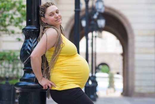 Smiling pregnant woman is leaning on a light pole outdoors during spring or summer time.