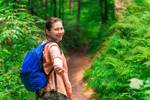 brunette with a backpack stretches her hand in a green forest in a hike