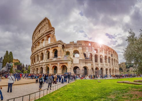 Rome, Italy, march 2017: tourists walking under the colosseum in rome on a cloudy day with the sun shining behind