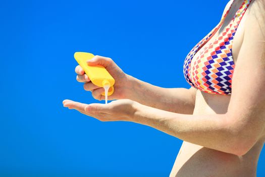 Closeup shot of female hands with a yellow bottle of sun protection cream on the blue sky background. Skin care and protection concept.