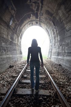 Man silhouetted in a tunnel standing in the center of the railway tracks looking towards the light at the end of the tunnel in a conceptual image