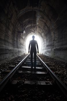 Man silhouetted in a tunnel standing in the center of the railway tracks looking towards the light at the end of the tunnel in a conceptual image
