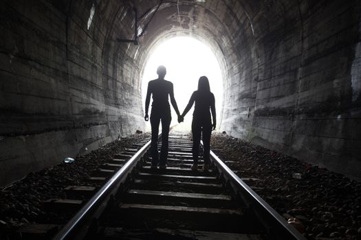 Couple walking hand in hand along the track through a railway tunnel towards the bright light at the other end, they appear as silhouettes against the light