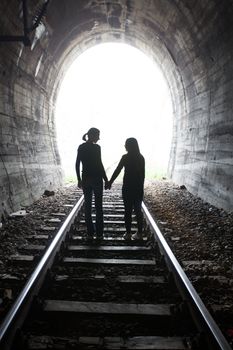 Couple walking hand in hand along the track through a railway tunnel towards the bright light at the other end, they appear as silhouettes against the light