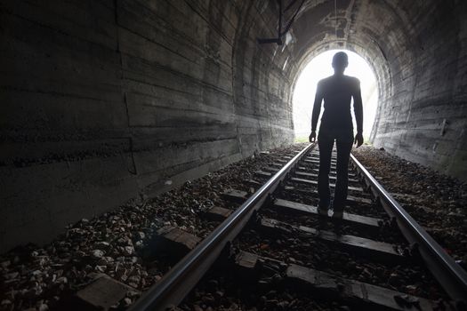 Man silhouetted in a tunnel standing in the center of the railway tracks looking towards the light at the end of the tunnel in a conceptual image