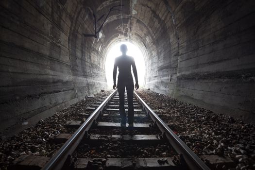 Man silhouetted in a tunnel standing in the center of the railway tracks looking towards the light at the end of the tunnel in a conceptual image