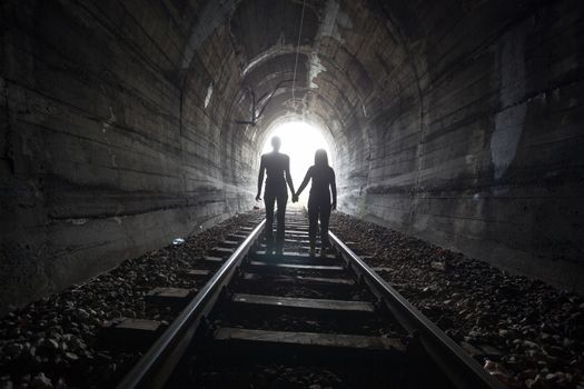 Couple walking hand in hand along the track through a railway tunnel towards the bright light at the other end, they appear as silhouettes against the light