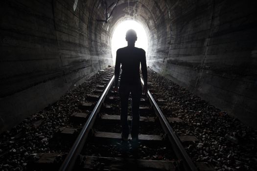 Man silhouetted in a tunnel standing in the center of the railway tracks looking towards the light at the end of the tunnel in a conceptual image