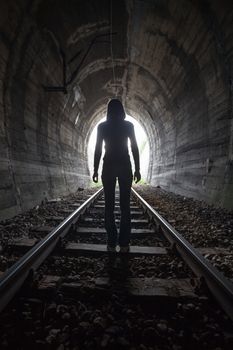 Man silhouetted in a tunnel standing in the center of the railway tracks looking towards the light at the end of the tunnel in a conceptual image