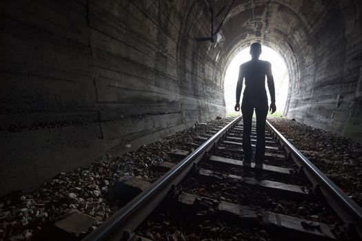 Man silhouetted in a tunnel standing in the center of the railway tracks looking towards the light at the end of the tunnel in a conceptual image