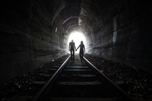 Couple walking hand in hand along the track through a railway tunnel towards the bright light at the other end, they appear as silhouettes against the light