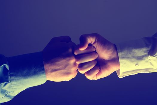 Side view with light source from below of fist bump handshake between two man over dark gray background