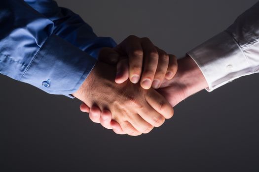 Close up side view of two male hands shaking over dark gray background and light source at bottom side