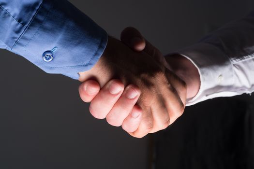 Low angle view of two businessmen hands shaking over dark gray background and light source at bottom side