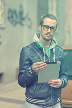 Young man with yellow glasses use iPad tablet computer on street, public space. Blurred background