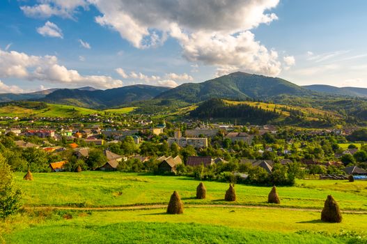 beautiful rural landscape in mountains. haystacks on the grassy hills. village at the foot of the mountain. interesting cloud formation over the ridge. lovely sunny afternoon