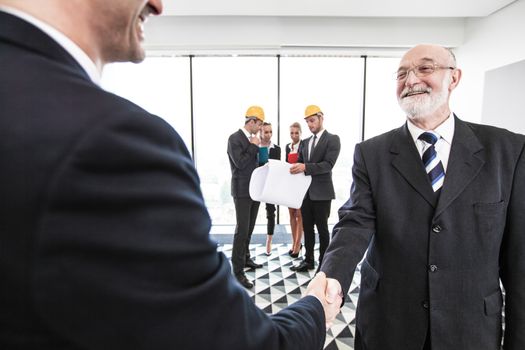 Business people at meeting shaking hands, their team in hardhats and blueprint on background, architects and investors