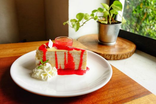 Crepe cake with cream and strawberry on top and sauce in ceramic cup on white plate.