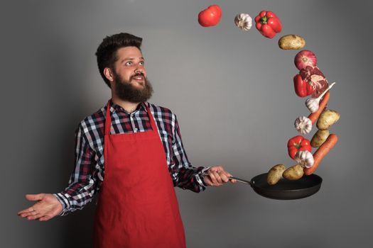 Cooking man concept, smiling bearded man in checked shirt, drop up meat and vegetables from a pan, studio shot on gray background