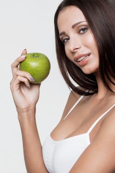 Side view of attractive brunette in sports bra biting green apple while standing on white background.