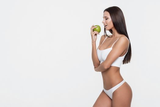 Side view of attractive brunette in sports bra biting green apple while standing on white background.