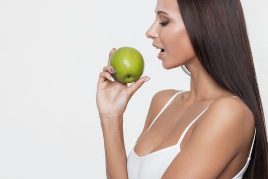 Side view of attractive brunette in sports bra biting green apple while standing on white background.