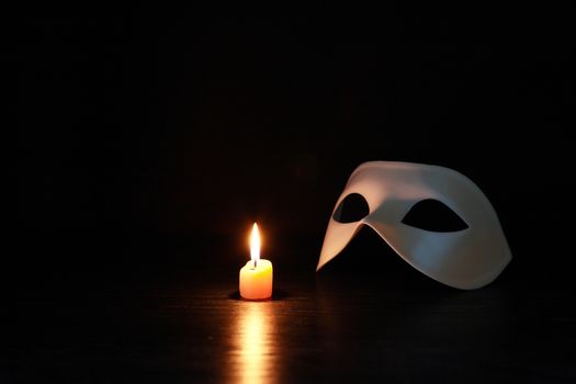 Classical white Venetian mask near lighting candle on dark background