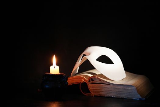 Classical white Venetian mask on old book near lighting candle against dark background