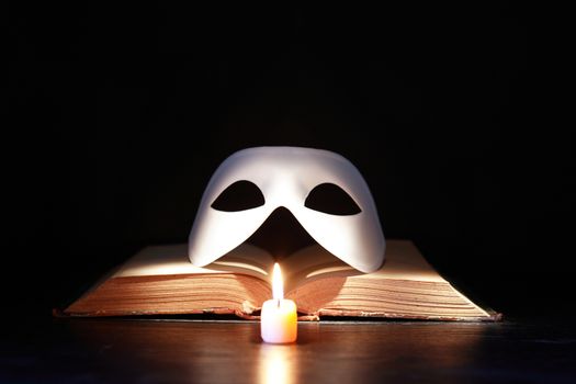 Classical white Venetian mask on old book near lighting candle against dark background