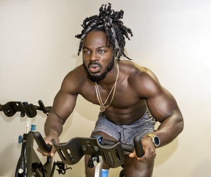 Handsome, young, muscular African American man, a bodybuilder, riding as stationary bicycle in the gym.