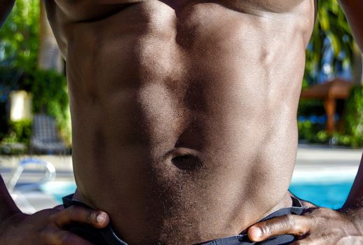 Torso of a young muscular handsome shirtless African American man showing his six pack, or abs, by the pool.