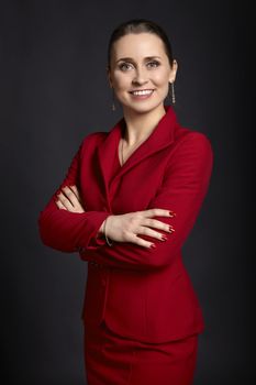 Portrait of elegant young business woman in red skirt suit with white smile and crossed arms, on black background.