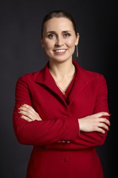 Portrait of elegant young business woman in red jacket with white smile and crossed arms, on black background.