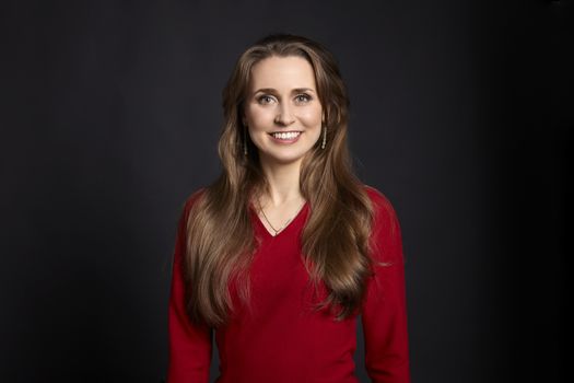 Studio portrait of young smiling woman in red dress with long hair, white smile and green eyes on black background.