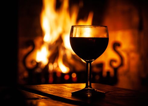 Glass of red wine on the wooden table with burning fire on the background. Evening relax on cozy place. Dark medieval style winery.
