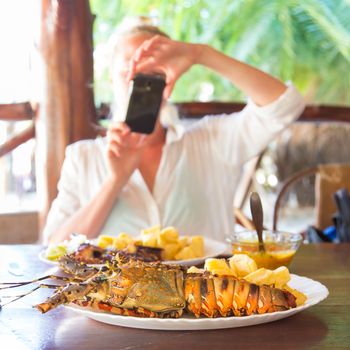 Foodie blogger on tropical vacations using mobile phone to take photo of a plate of grilled lobster served with potatoes and coconut sauce.