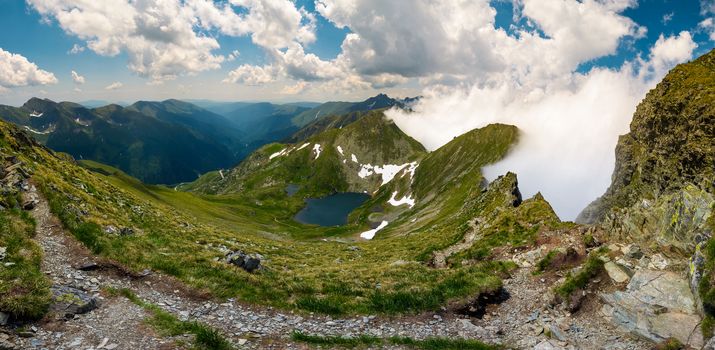 Gorgeous landscape of Fagaras mountains in summer. clouds rising above the rocky cliffs. lake Capra down the grassy slope in the valley. view from the tourist path on top of the ridge.