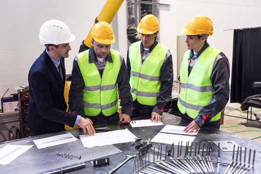 Engineer, foreman and workers wearing safety helmets are meeting at factory and discussing papers and bluprints at the table
