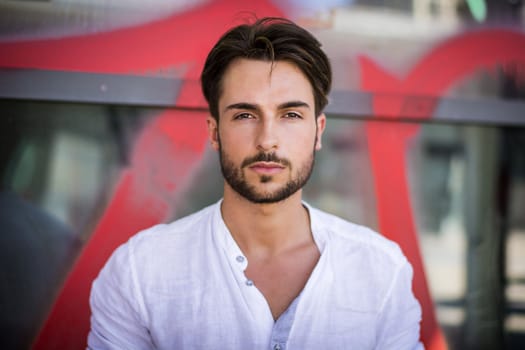 Stylish trendy young man standing outdoor against office window, looking confindent at camera