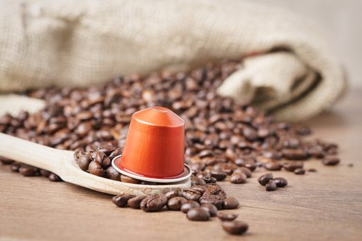 In the foreground a coffee capsule on wooden spoon and   roasted coffee beans with burlap sack on blur wooden background ,close up.